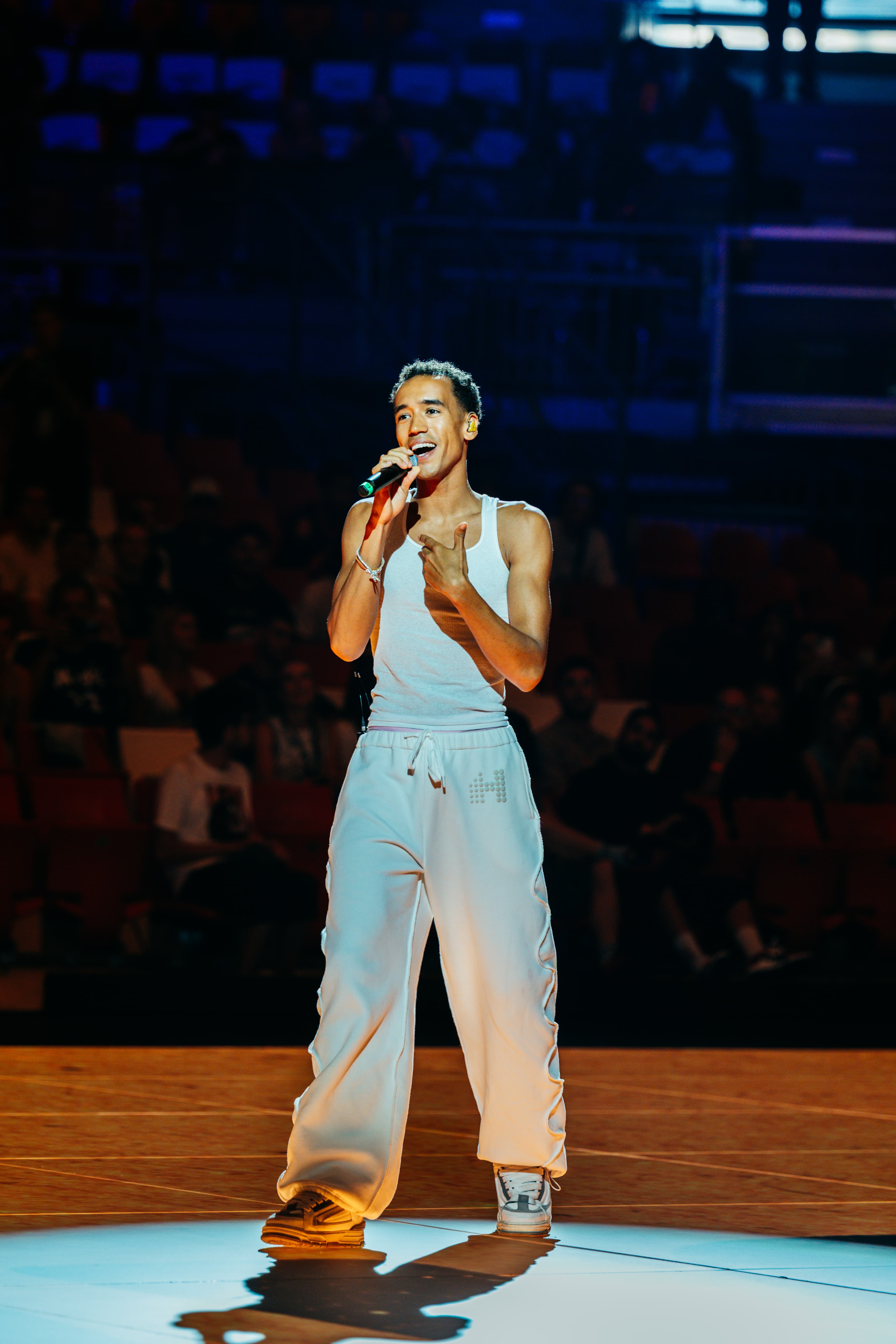 WizTheMc in a white tank top smiling while singing into a mic under a spotlight on the court