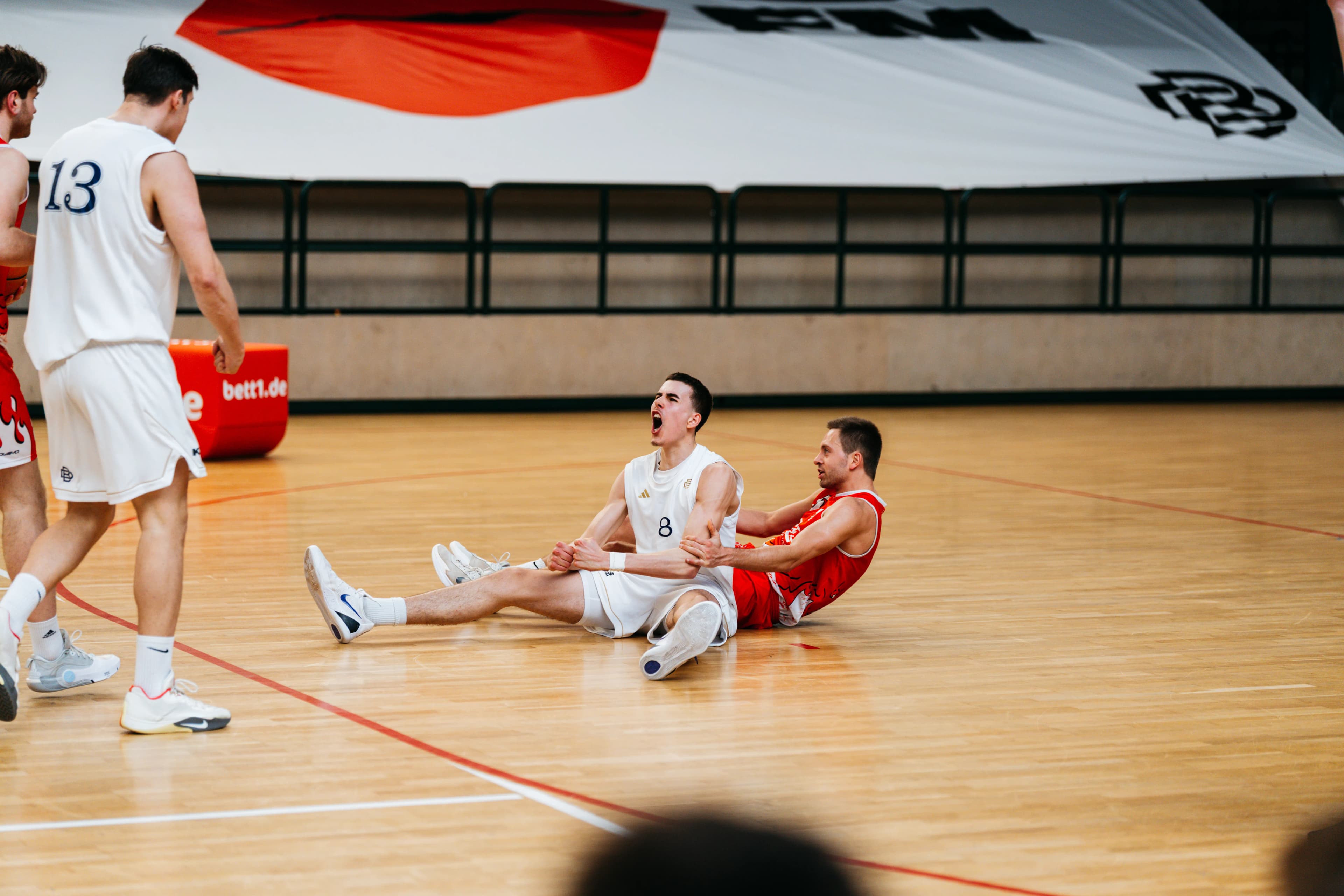 Berlin Dreams player number 8 sitting on the court shouting in celebration, opponent in red behind, Bett1 sideline pad