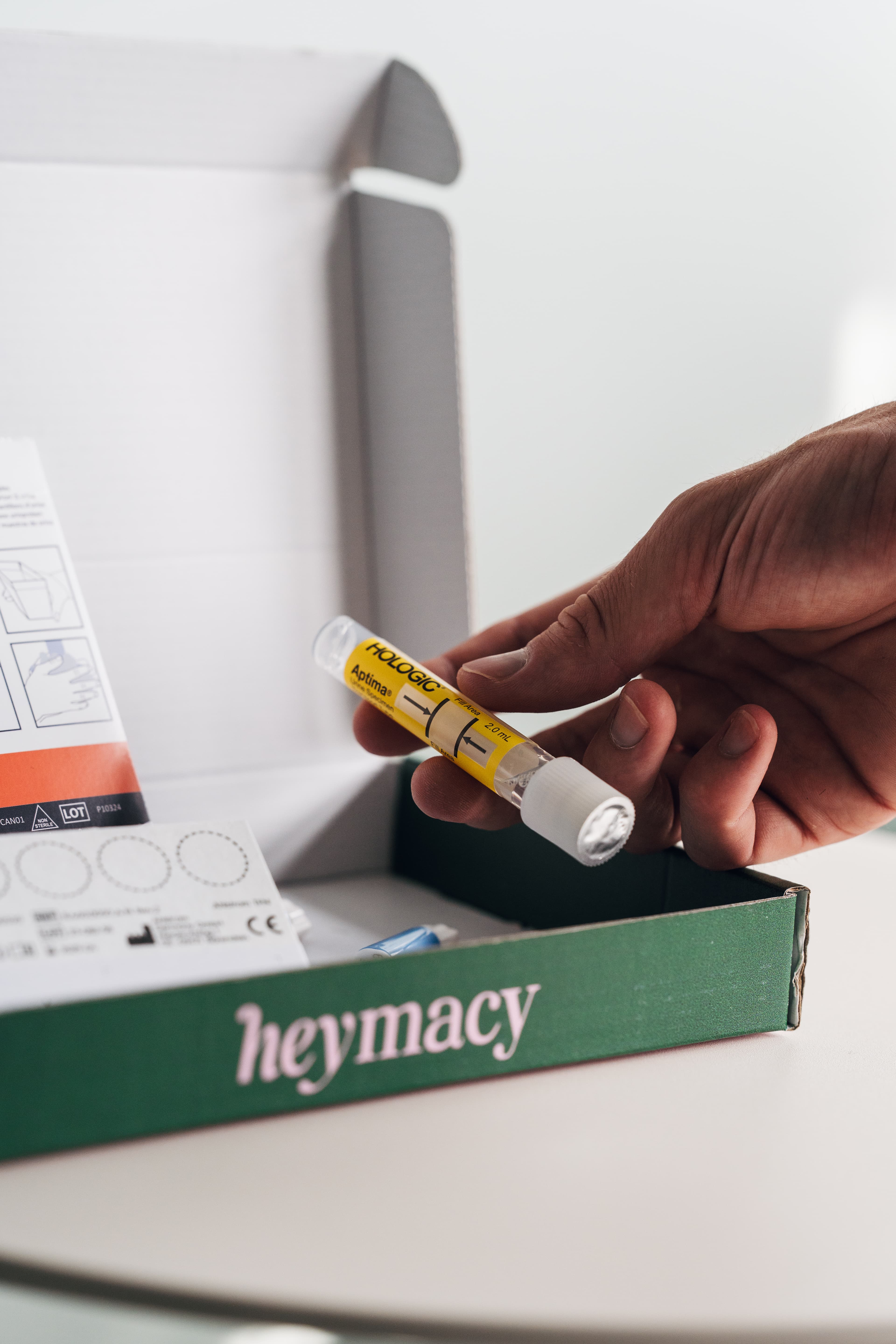 Hand holding a clear test tube with a yellow label above the open heymacy box
