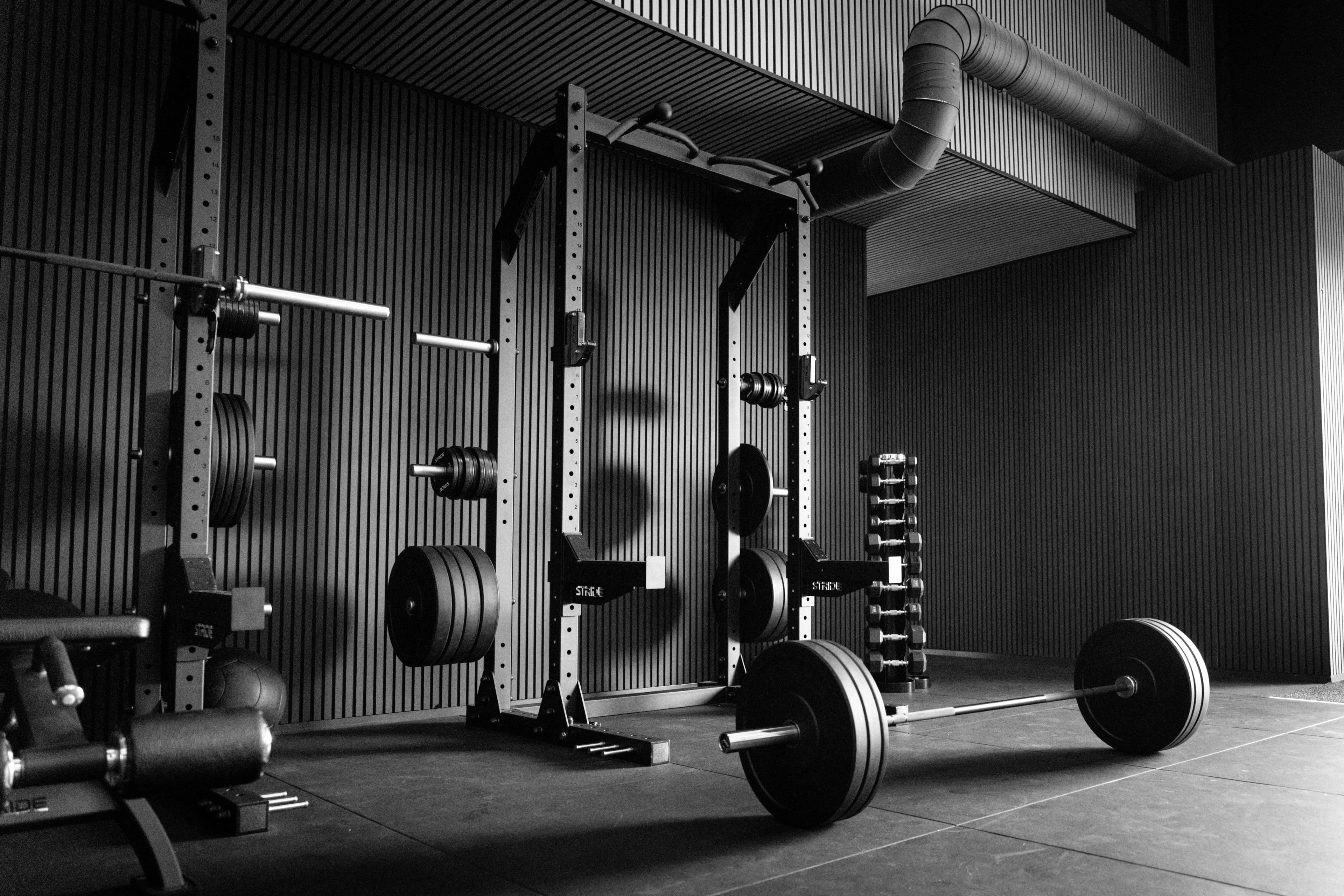 Black and white: barbell with bumper plates in front of power racks and corrugated metal walls in the Kraftwerk Neuhof training room