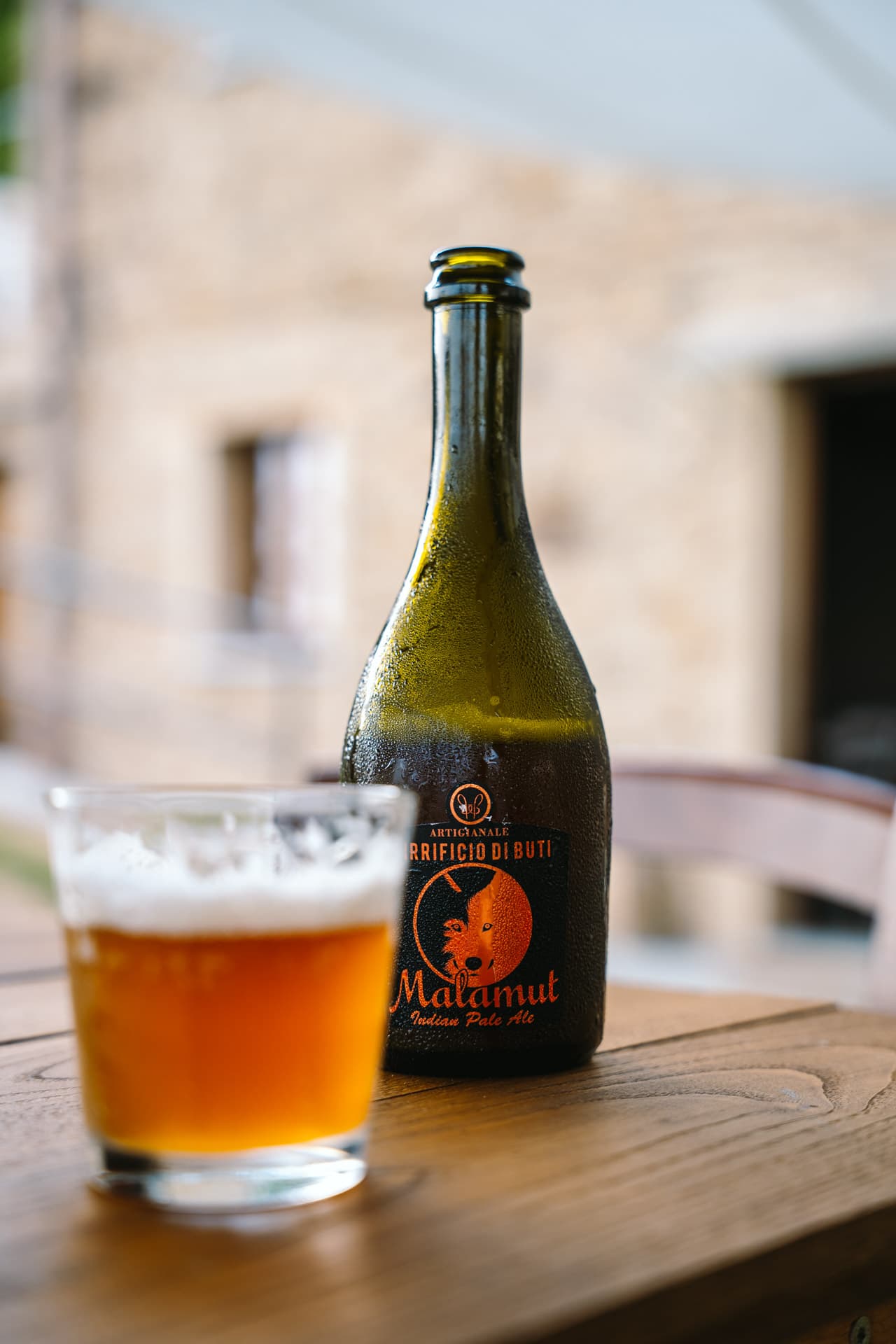 Condensation-covered Malamut IPA bottle and frothy glass on a wooden table against a stone façade