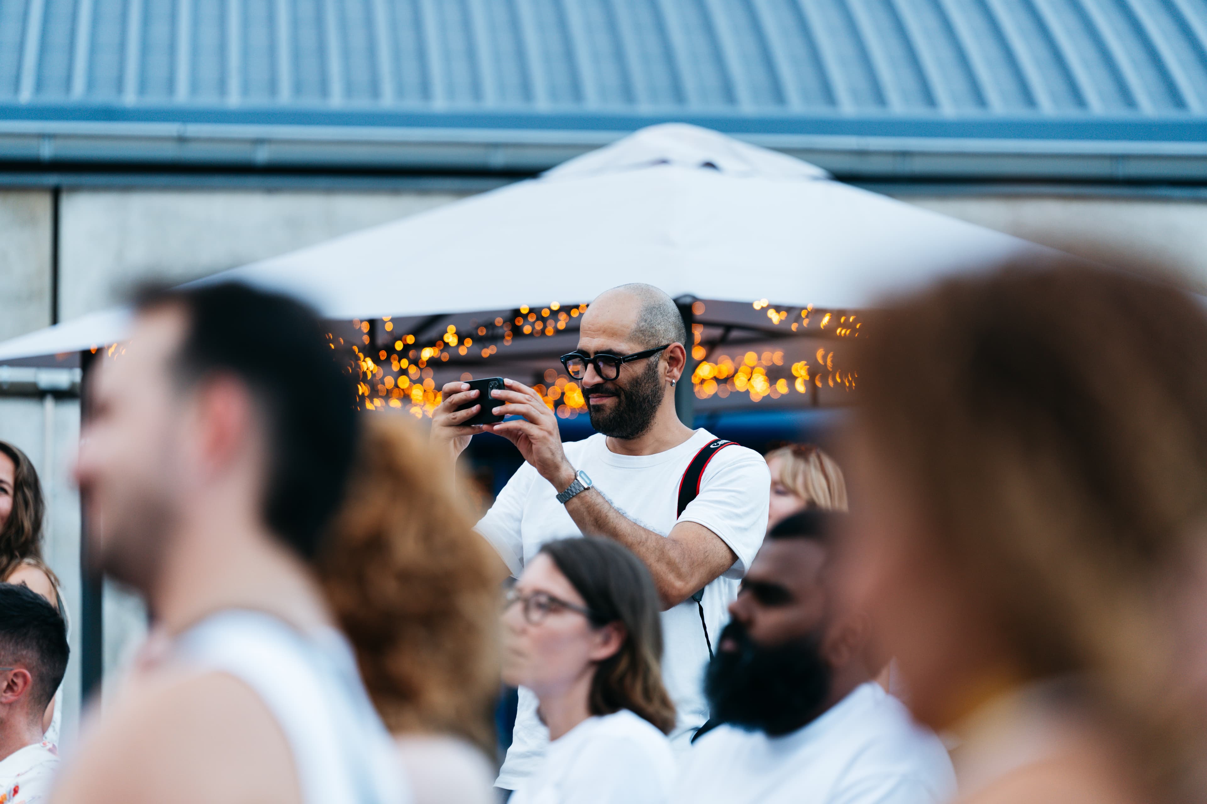 Guest with glasses and a white shirt smiling while filming on a smartphone under string lights